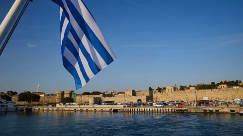 Departure of a ferry, Greek flag in front of harbour and historic city walls under blue sky,