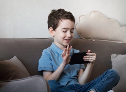 A young boy is sitting on a couch, happily engaging with a smartphone. He wears a blue shirt and