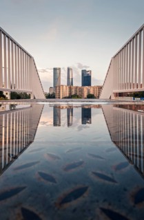 A stunning view of Madrid's skyscrapers reflected in a puddle on a bridge at sunset. The serene sky