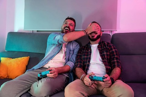A gay couple sitting on a couch, joyfully playing video games. One partner covers the other's eyes,