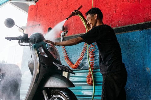 A professional motorcycle cleaner uses a pressure washer to thoroughly clean a scooter. The vibrant