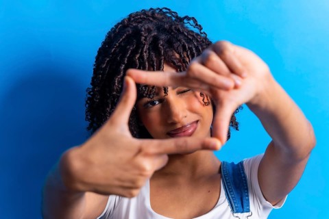 Young woman with curly hair making a frame with her hands around her face, winking, on a blue