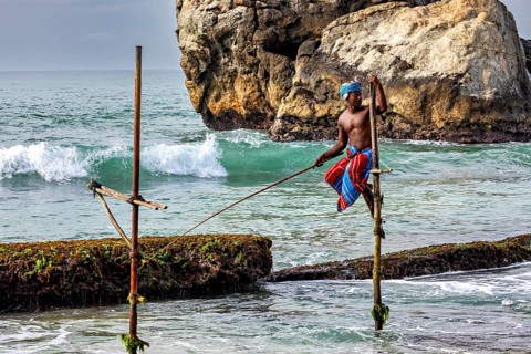 A fisherman balances on a stick above the waves off a rocky coast, The stilt fishermen on the beach