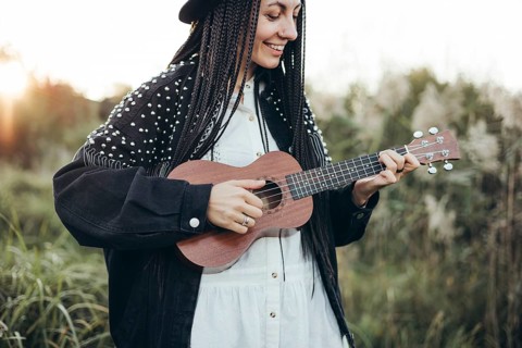 A woman with braids smiles while playing a ukulele at sunset in a serene green landscape. Her