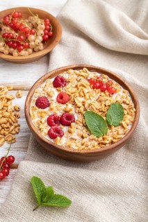 Wheat flakes porridge with milk, raspberry and currant in wooden bowl on white wooden background