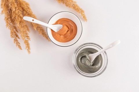 Top view of two bowls of cosmetic clay facial masks, one green and one orange, with white spoons