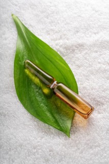 Glass ampoule of anti-aging serum on a fresh green leaf, placed on a soft white towel. Ideal for