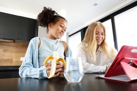 A smiling girl eating a banana and a young woman share a joyful moment in a bright, modern kitchen.