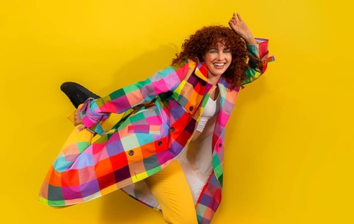 A joyful young woman leaps against a bright yellow backdrop, wearing a multicolored checkered coat.