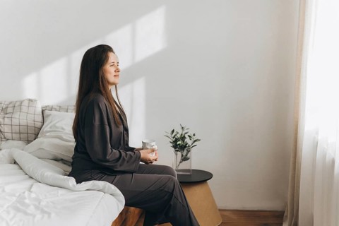 A woman sits on a bed, holding a cup, while gazing out the window in a bright, calming room. The