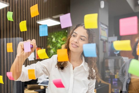 A woman in a modern office uses colorful sticky notes on a glass wall for planning. She's focused