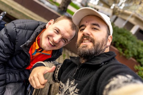 Young man in a wheelchair and his friend are smiling and giving a thumbs up while taking a selfie