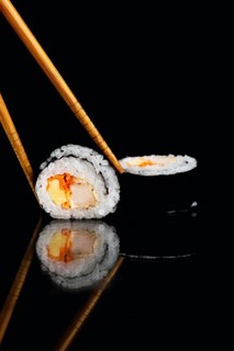 Close up of a sushi roll held by chopsticks with a reflection on a sleek black surface. The vibrant