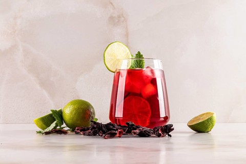 A glass of refreshing red hibiscus tea with ice cubes, garnished with a lime slice and mint leaves.