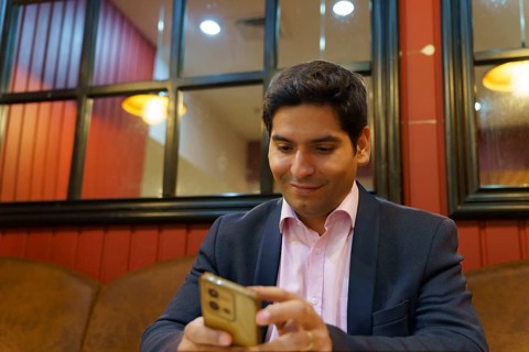 A young Chilean businessman in a suit, seated in a cozy cafe, engrossed in his smartphone. The warm