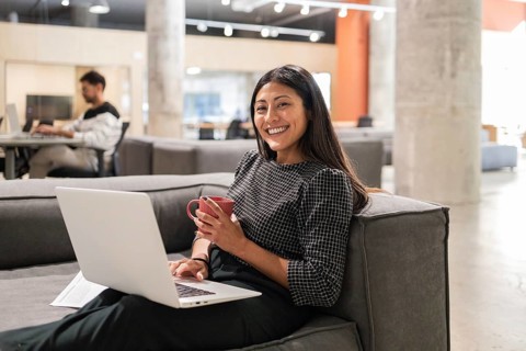 A young professional woman smiles while working on a laptop, holding a coffee mug in a modern