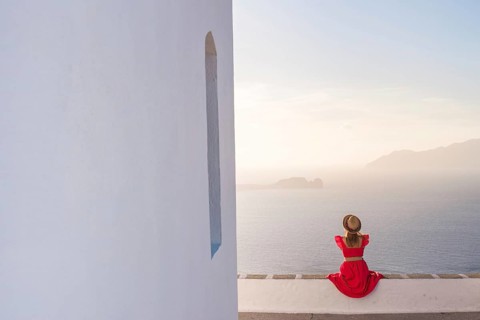 A woman in a red dress and hat sits on a white wall, overlooking a serene ocean and distant