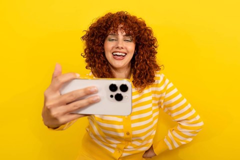 A smiling woman with curly red hair in a yellow striped shirt takes a selfie against a vibrant