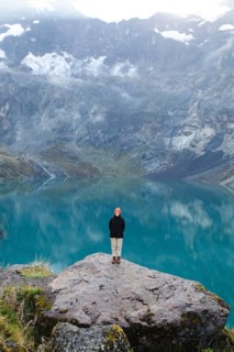 An Ecuadorian woman in a hat and jacket stands on a rock overlooking the turquoise lake at El Altar
