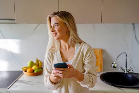 A woman with long blonde hair smiles while holding a phone in a modern kitchen. She wears a cozy