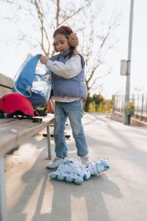 A young girl on roller skates is organizing her belongings in a blue bag at a park bench, ready for