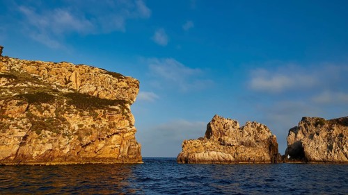Cliffs with distinctive shapes rise out of the calm blue sea under a cloudy sky, morning light,
