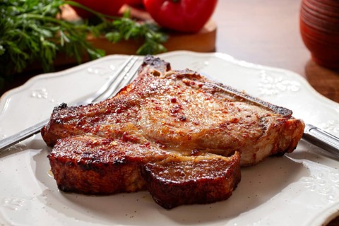 A pork chop rests on a white plate with a fork and knife. Fresh tomatoes and green herbs surround