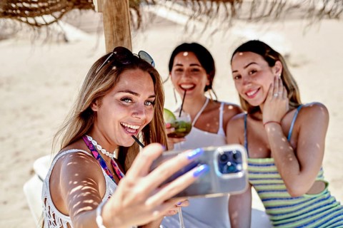 Three friends gather under a straw umbrella on the beach, joyfully taking a selfie and sipping