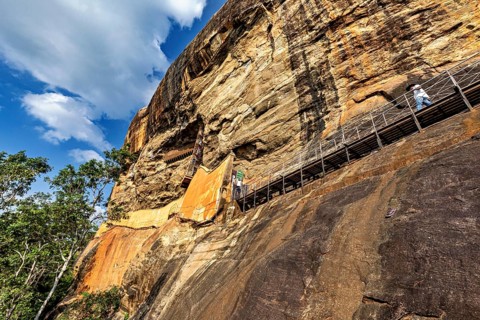 A narrow path leads along an imposing rock face over which a blue sky arches, The Lion Rock near
