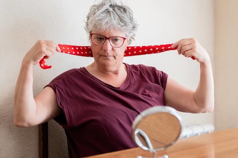 A senior woman with short grey hair and glasses ties a red polka dot scarf around her neck. She