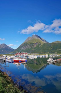 Wooden houses, small fishing boats and steep mountains reflected in the water of a harbour,