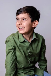 A young boy with dark hair, wearing a green button-up shirt, smiles broadly while looking to the