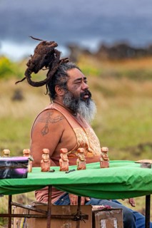 A man with dreadlocks and beard sits outside at a table with small statuettes in a meadow,