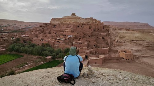 A man sits on a hill and looks at Ait-Ben-Haddou a historic clay town and the vast countryside,