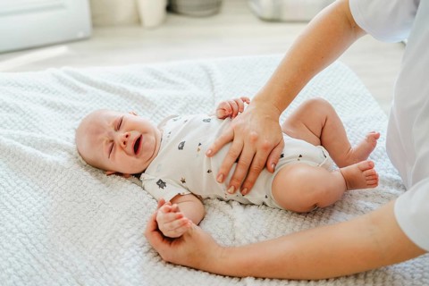 A mother comforts a crying baby lying on a soft white blanket. The infant wears a cute, patterned