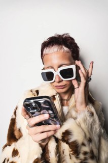 Stylish woman in a faux fur coat, large sunglasses, and jewelry, focused on a smartphone. The image