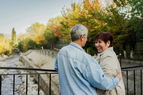 A joyful elderly couple shares a tender moment on a riverside bridge amidst vibrant autumn foliage.