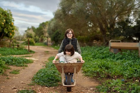 Two children playfully enjoy a ride in a wheelbarrow on a scenic rural path surrounded by lush