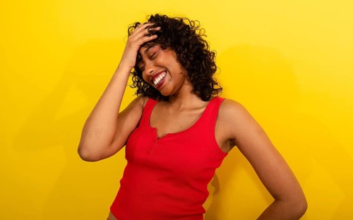 A woman in a vibrant red tank top laughs joyfully, hand on her head, against a bright yellow