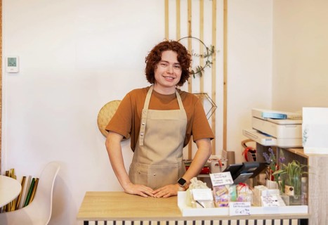 A young man wearing an apron stands smiling behind a organic products shop counter The warm and
