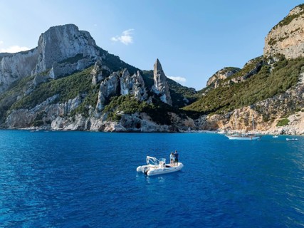Couple on motorboat off picturesque rocky coast, cliffs with L'Aguglia pinnacle, blue sea and Cala