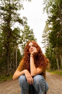 A young woman with red hair enjoys a peaceful moment in a Swedish forest, surrounded by tall trees.