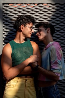 A joyful gay couple, wearing colorful outfits and sunglasses, shares a tender moment under the warm