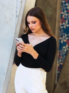 A woman in a black top and white pants leans against a concrete wall while using a smartphone. She