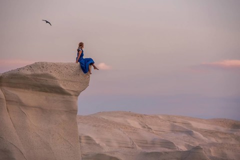 Unrecognizable woman wearing a blue dress perched on a high white rock formation at Sarakiniko on