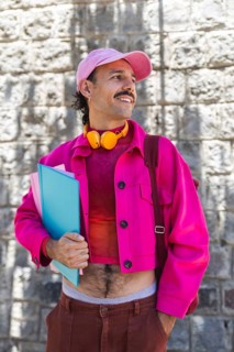 A young Spanish man with a mustache, dressed in vibrant pink clothing, holds folders while enjoying