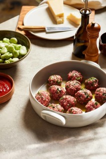 Raw meatballs seasoned with herbs in a white dish ready for cooking, surrounded by fresh