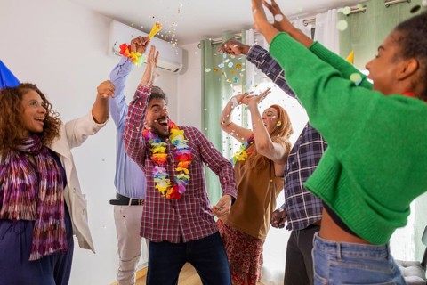 A lively group of friends celebrates a birthday indoors with laughter, party hats, colorful leis,