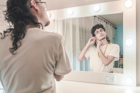 Young man with curly hair and glasses adjusting his earring while gazing into a mirror in a cozy