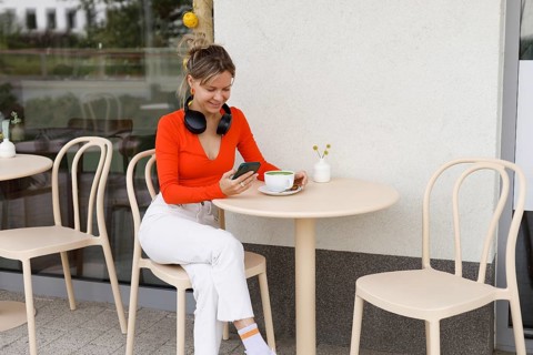 A woman sitting at an outdoor cafe table in Warsaw, wearing headphones and using a smartphone She